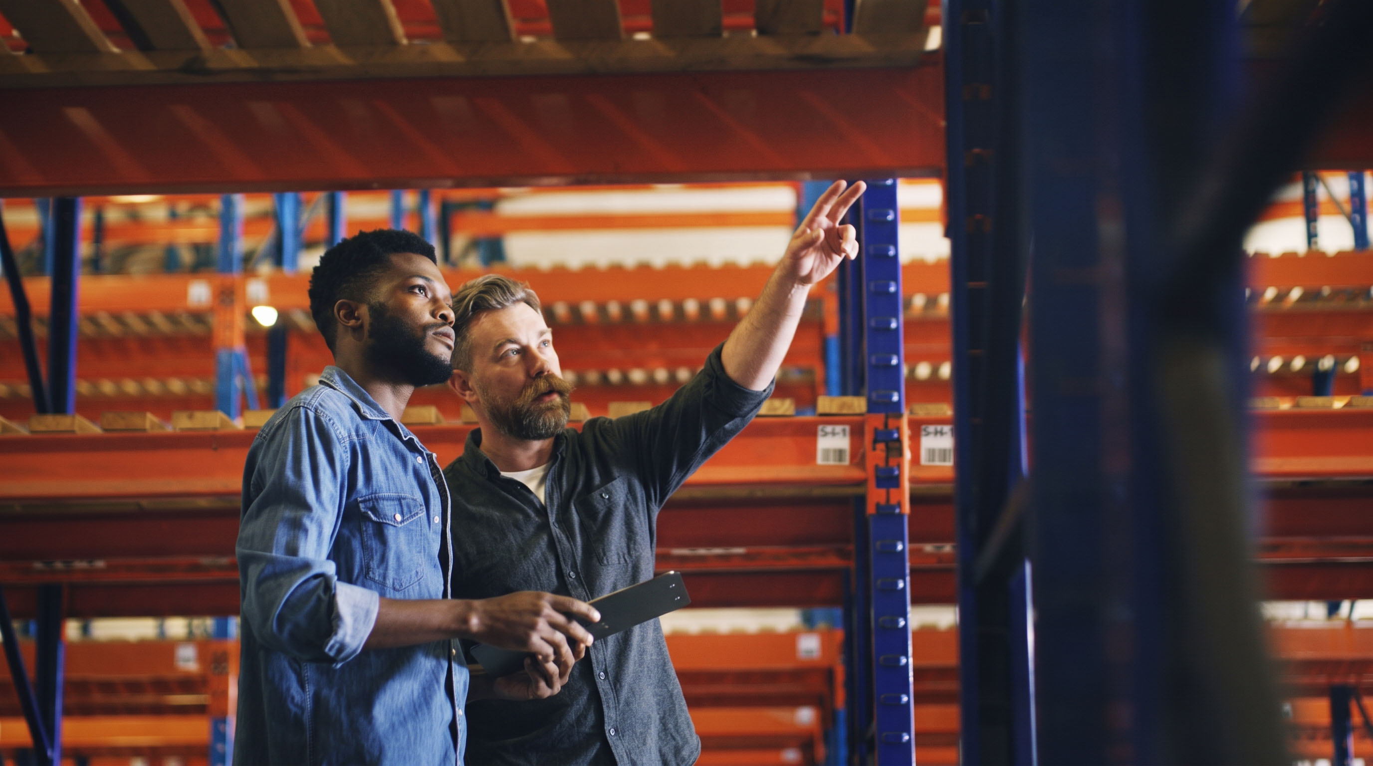 Two men in a warehouse discuss inventory while one points and the other holds a clipboard.
