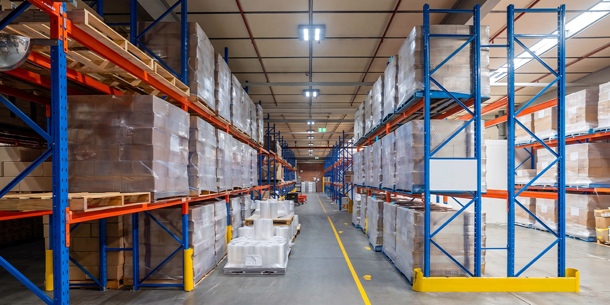 Emrack pallet racking for Ross Labs. Warehouse aisle with tall blue and orange metal racks filled with pallets of wrapped boxes and packages.