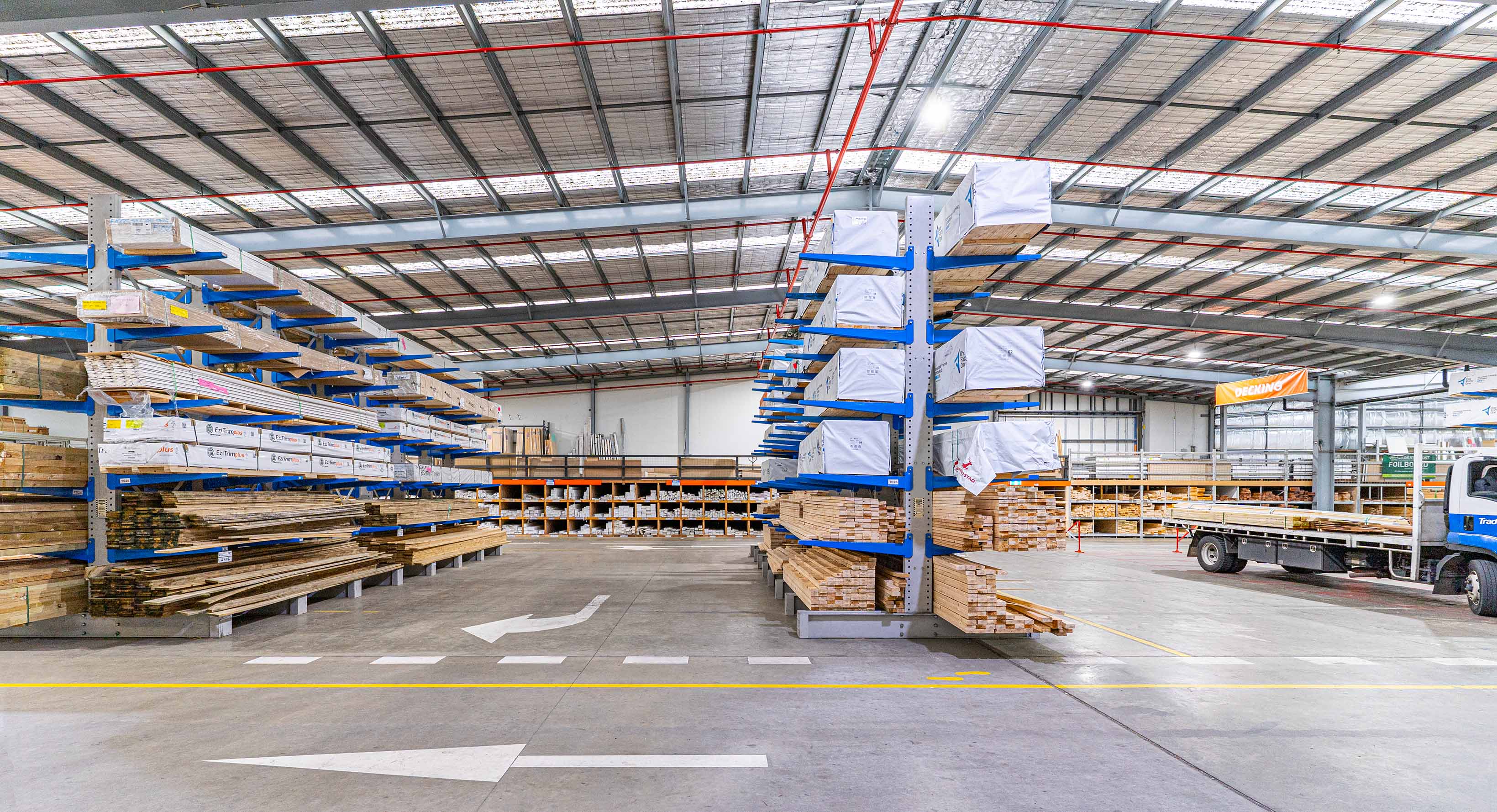 Indoor warehouse of stacked timber boards on cantilever racking and a truck on the right side under a metal roof.
