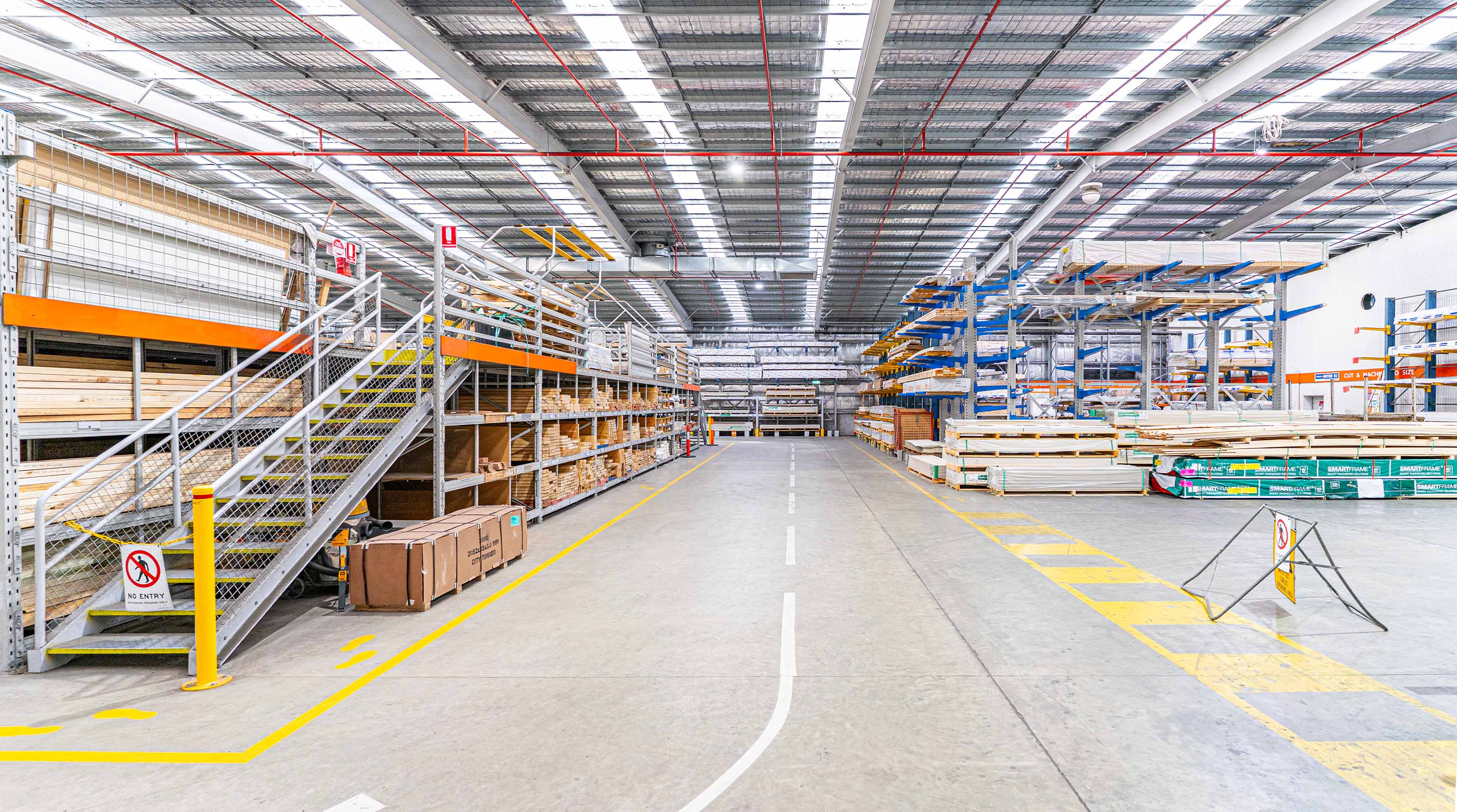 Wide warehouse aisle with cantilever racking, pigeon hole and mezzanine racking under a high ceiling with exposed pipes.