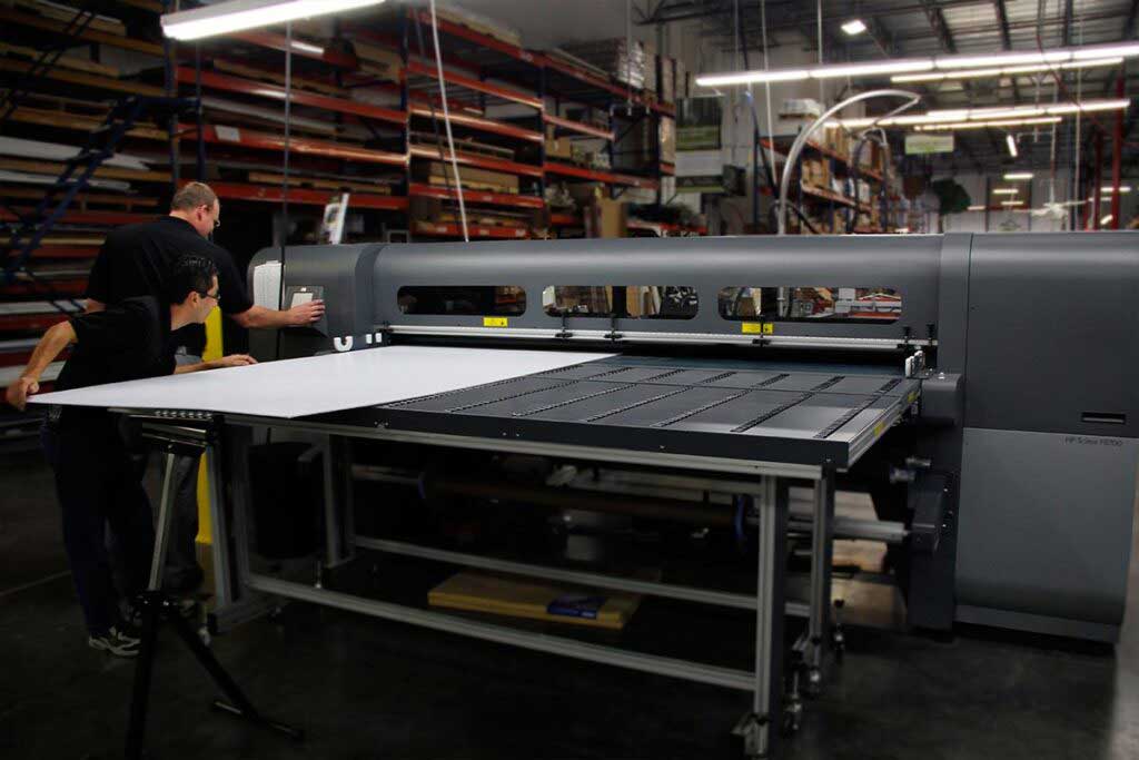 Two men operating a large industrial printing machine in a warehouse filled with shelves.