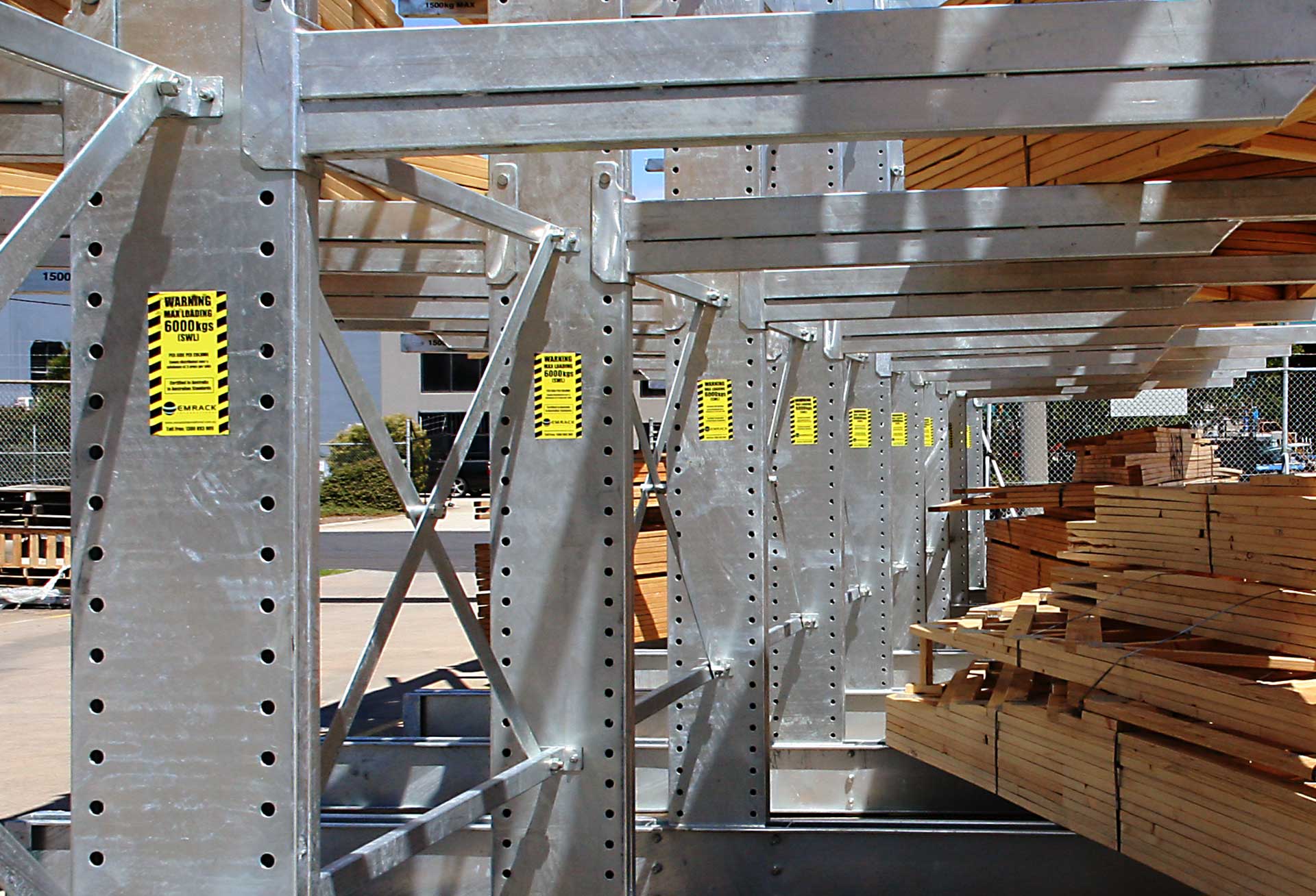 Metal racking system with yellow warning labels indicating maximum load of 6000 kgs, storing bundles of lumber outside.